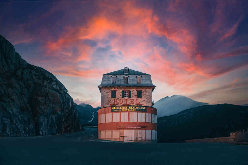 Solitary hotel on the inside curve of a winding mountain road, below a spectacular late sky of wispy red-hued clouds and with mountains behind in the distance and to the sides.